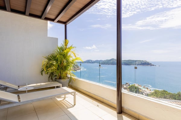 Wooden deck with lounge chairs and black umbrella overlooking turquoise bay, forested hills, and glass railing