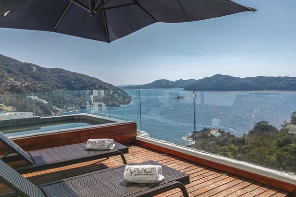 Wooden deck with lounge chairs and black umbrella overlooking turquoise bay, forested hills, and distant islands