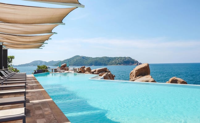 Covered terrace with wooden table and chairs overlooking turquoise sea, rocky coastline, and distant island